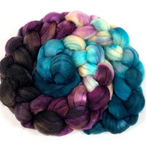 May include: A braided bundle of colorful wool roving in shades of teal, purple, and cream. The wool is arranged in a circular pattern, with the colors blending together. The texture of the wool is soft and fluffy, perfect for fiber art projects.