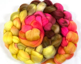 Può includere: Un pacchetto di lana cardata intrecciata in diversi colori, dal giallo all'arancione, passando per il rosa e il marrone. La lana è disposta in forma circolare, con le estremità delle trecce che si sovrappongono.