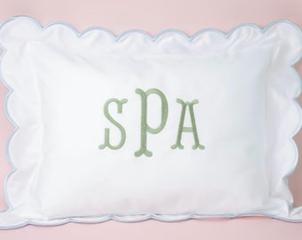 Puede incluir: Almohada rectangular blanca con borde festoneado y ribete azul claro. La almohada presenta la palabra "SPA" bordada en verde. El fondo es de color rosa claro liso.