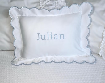 Puede incluir: Almohada blanca con borde festoneado y ribete azul claro. La palabra "Julian" está bordada en azul claro en la funda de almohada. La almohada está sobre una colcha blanca.
