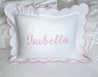 Puede incluir: Almohada rectangular blanca con un borde festoneado rosa y el nombre "Isabella" bordado en escritura rosa. La almohada está sobre una cama blanca con otras almohadas blancas.