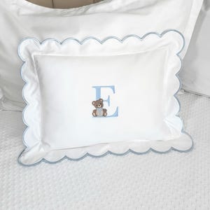 Puede incluir: Almohada rectangular blanca con borde festoneado azul claro. La almohada presenta una letra "E" bordada en azul claro con un pequeño diseño de oso de peluche. La almohada está sobre una cama blanca con un patrón texturizado.