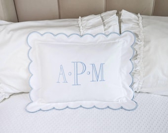 Puede incluir: Almohada rectangular blanca con borde festoneado azul claro e iniciales bordadas "A.P.M." en azul claro. La almohada está sobre una cama blanca con fundas de almohada con volantes.