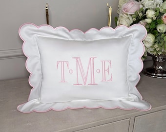 Puede incluir: Almohada rectangular blanca con borde festoneado rosa e iniciales rosas bordadas "TME". La almohada está sobre una superficie clara, con un arreglo floral en el fondo.