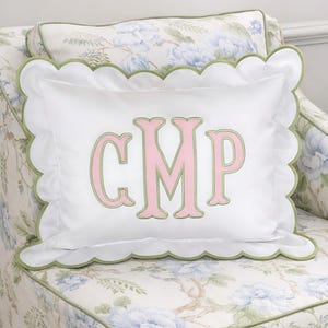 Op de afbeelding: Wit decoratief kussen met een geschulpte rand en groene bies. Het kussen heeft de geborduurde initialen "CMP" in roze met een groene omtrek. Het kussen ligt op een stoel met bloemenpatroon.