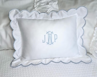 Puede incluir: Almohada rectangular blanca con borde festoneado y ribete azul claro. La almohada presenta un monograma bordado en azul claro. La almohada está sobre una cama blanca con una funda de almohada con volantes.