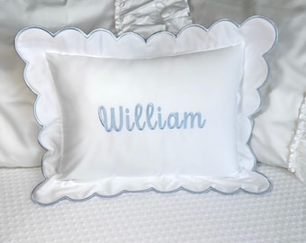 Puede incluir: Almohada rectangular blanca con borde festoneado, ribeteada en azul claro. El nombre "William" está bordado en escritura azul claro en el centro. La almohada está sobre una superficie blanca texturizada.