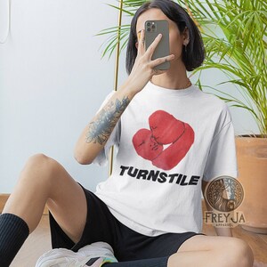 May include: White t-shirt with a red graphic of two boxing gloves in a heart shape. The text "TURNSTILE" is printed in black below the graphic.