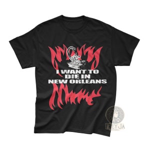 Pode incluir: Camiseta preta com um design gráfico vermelho que diz "Eu quero morrer em Nova Orleans". O design gráfico apresenta um escorpião estilizado e chamas.