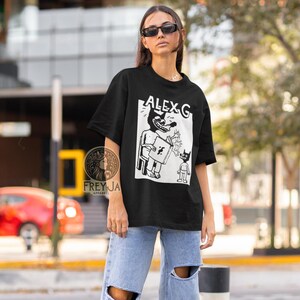 Op de afbeelding: Zwart T-shirt met een witte grafische print van een cartoonfiguur dat een boek vasthoudt en een ander kleiner figuur. De tekst "ALEX G" is op het shirt gedrukt.