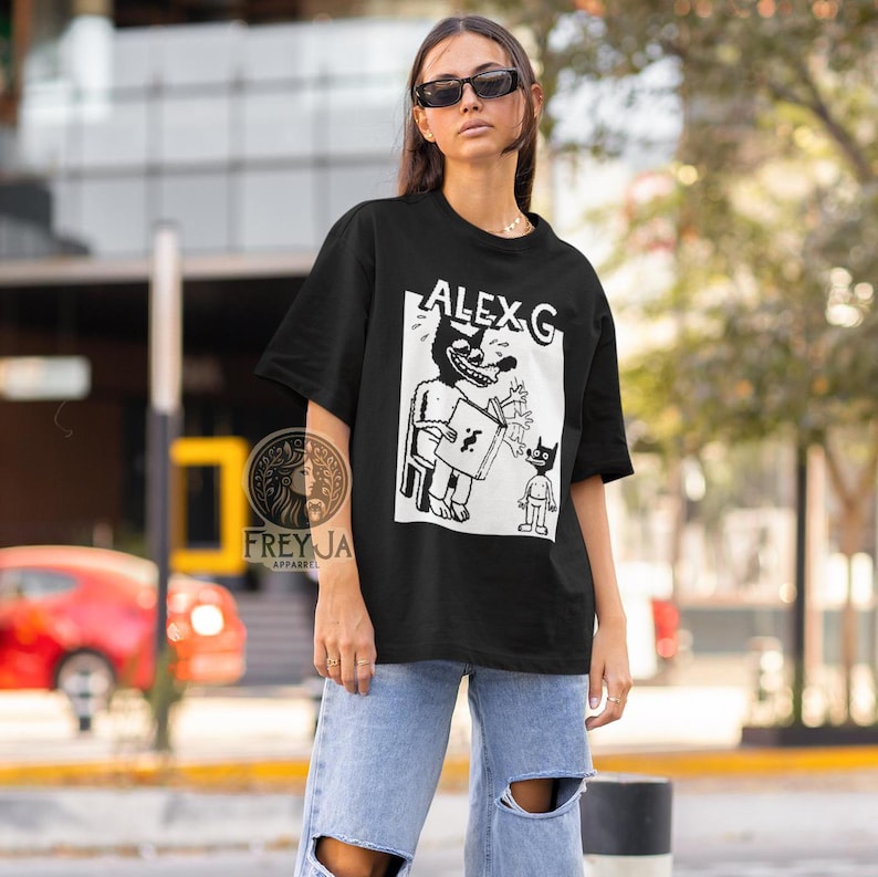Op de afbeelding: Zwart T-shirt met een witte grafische print van een cartoonfiguur dat een boek vasthoudt en een ander kleiner figuur. De tekst "ALEX G" is op het shirt gedrukt.