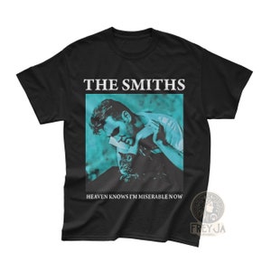 Puede incluir: Camiseta negra con una impresión blanca de una banda llamada "The Smiths". La impresión presenta una foto de un hombre con un gato en un esquema de color azul y verde. El texto "Heaven Knows I'm Miserable Now" está impreso debajo de la foto.