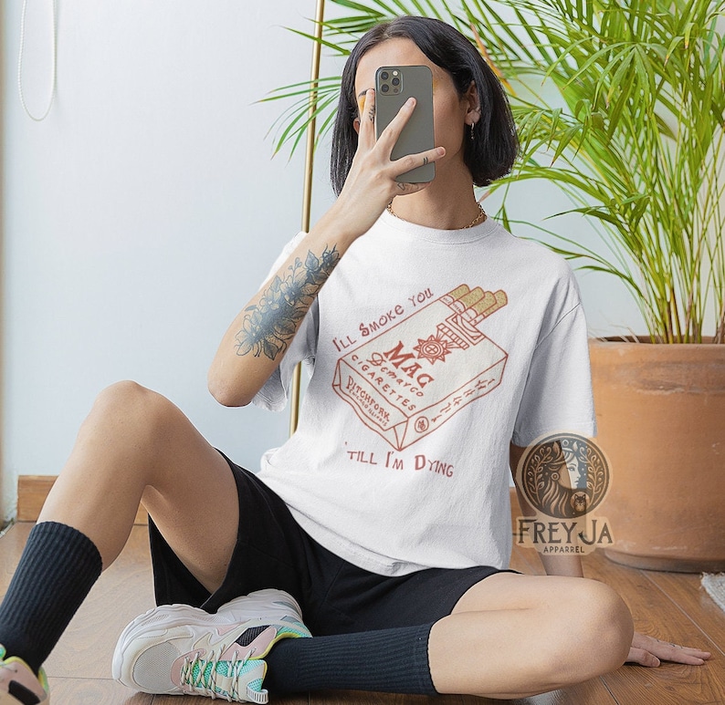May include: White t-shirt with a graphic of a vintage cigarette pack that says "Mag Demarco Cigarettes Pitchfork" and the text "I'll smoke you 'till I'm dying".
