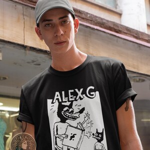 Op de afbeelding: Een zwart T-shirt met een witte grafische print van een cartoonfiguur met de tekst "ALEX G" erboven.