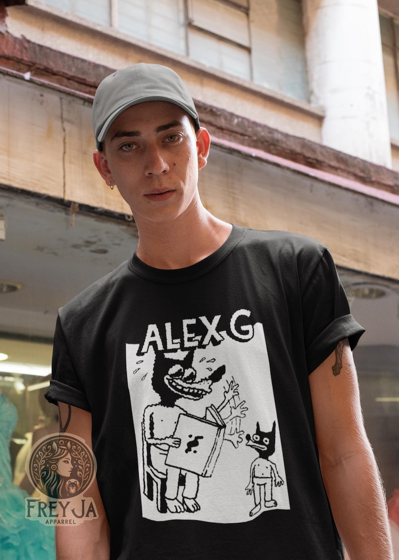 Op de afbeelding: Een zwart T-shirt met een witte grafische print van een cartoonfiguur met de tekst "ALEX G" erboven.