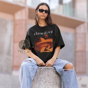 以下が含まれることがあります： 前面に白い文字で「slowdive」というバンド名がプリントされた黒いTシャツ。シャツには、バンドのアルバム「Just for a Day」のカバーアートがオレンジとイエローの暖色系で描かれています。