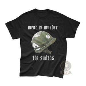 Puede incluir: Camiseta negra con texto blanco que dice "meat is murder the smiths". La camiseta presenta un casco militar verde con una pegatina blanca en el lateral.