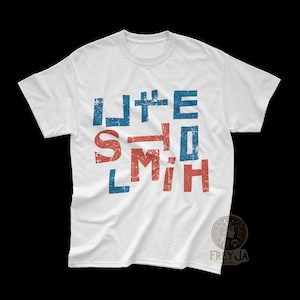 Op de afbeelding: Wit t-shirt met de tekst "LITE HOLMITH" in een verweerde, blokkerige letter. De letters zijn blauw en rood. Het shirt is gemaakt van een zacht materiaal.