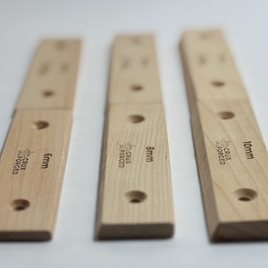 May include: Three light-colored wooden climbing holds with rounded edges. Each hold has a hole and the text "CRUX FORGED". The holds are labeled 6mm, 8mm, and 10mm. The holds are for rock climbing.