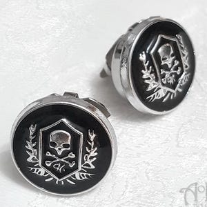 May include: A pair of silver skull and crossbones cufflinks with black enamel. The skull is inside a shield with a wreath of leaves around it.