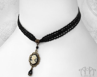 Op de afbeelding: Zwarte kanten choker ketting met een camee hanger. De hanger heeft een wit profiel tegen een zwarte achtergrond, omlijst in een bronskleurige setting. Een zwarte druppelvormige kraal hangt eronder. De ketting wordt getoond op een witte mannequin.