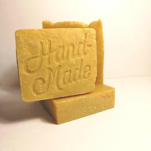 May include: Close-up of several square, light-yellow bars of soap. The top bar is imprinted with the words "Hand Made" in a cursive font. The soap bars are stacked, with one bar partially obscuring the others. The background is a plain white.