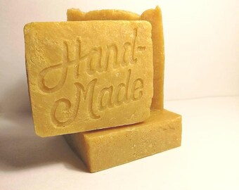 May include: Close-up of several square, light-yellow bars of soap. The top bar is imprinted with the words "Hand Made" in a cursive font. The soap bars are stacked, with one bar partially obscuring the others. The background is a plain white.