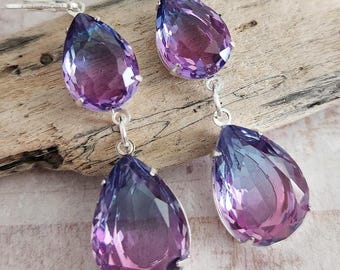 May include: A pair of silver-toned drop earrings featuring large, teardrop-shaped purple and blue gradient gemstones. The earrings are displayed on a piece of driftwood against a soft, patterned background.