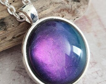 May include: A silver-toned necklace featuring a round pendant with a swirling design in shades of purple and blue. The pendant is set in a silver bezel and hangs from a silver chain. The necklace is displayed on a piece of driftwood.