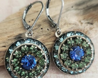 May include: A pair of silver-toned leverback earrings. Each earring features a circular design with a central blue stone surrounded by green and blue crystals. The earrings are set against a wooden background.
