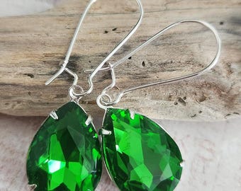 May include: A pair of emerald green teardrop earrings with silver-colored ear wires. The faceted green stones are set in a silver-colored metal. The earrings are displayed on a piece of driftwood.