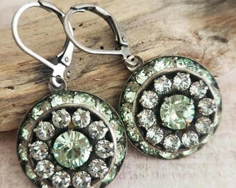 May include: A pair of silver-toned leverback earrings. Each earring features a circular design with a central light green stone surrounded by smaller clear stones. The outer edge is lined with more light green stones.