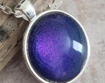 May include: A silver-toned necklace featuring a round pendant with a deep purple, iridescent center. The pendant is set in a silver bezel and hangs from a delicate chain. The purple center has a shimmering, galaxy-like appearance.