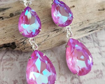 May include: A pair of dangling earrings with iridescent, teardrop-shaped crystals in shades of pink and purple. The crystals are set in silver-colored metal and are connected by small chains. The earrings are displayed on a piece of driftwood.