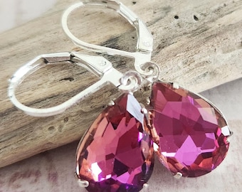 May include: A pair of silver leverback earrings with large, teardrop-shaped, faceted, pink and purple crystals. The earrings are displayed on a piece of driftwood.