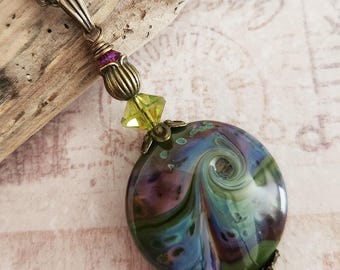 May include: A handcrafted necklace featuring a large, round pendant with swirling patterns of green, blue, and purple. The pendant is accented with brass-toned metal and colorful beads. The chain is also brass-toned.