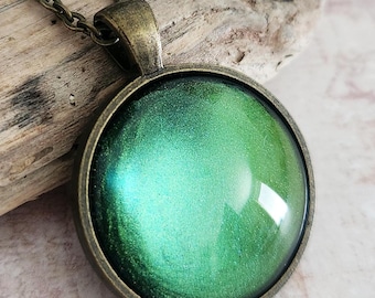 May include: A round pendant necklace with a bronze-colored chain and setting. The pendant features a vibrant, iridescent green center, creating a shimmering effect. The necklace is a piece of jewelry.
