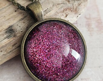 May include: A round pendant necklace with a bronze-colored chain and setting. The pendant features a sparkling, glittery, deep pink design. The necklace is displayed on a piece of driftwood.