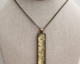 May include: A rectangular pendant necklace with a bronze-colored chain. The pendant has a bronze frame and contains colorful, glittery material. The necklace is displayed on a mannequin.
