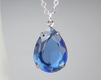 May include: A silver chain necklace with a large, teardrop-shaped, faceted blue pendant. The pendant is a deep sapphire blue color and is held in place by small silver prongs.