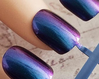 May include: A set of iridescent, oval-shaped false nails in shades of blue, purple, and pink. The nails are attached to a blue plastic holder, with one nail applied to a mannequin finger. The nails have a glossy finish.