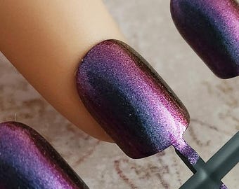 May include: A set of iridescent press-on nails in shades of purple, blue, and pink. The nails are oval-shaped and have a glossy finish. They are attached to a dark green plastic holder, with one nail applied to a finger.