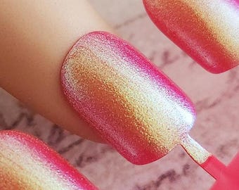 May include: Close-up of a set of iridescent press-on nails. The nails are oval-shaped with a pink and gold color gradient. They are attached to a pink plastic frame, ready for application. The nails are on a light-colored surface.