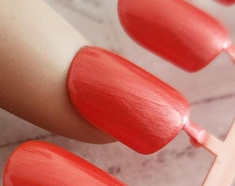 May include: Close-up of coral-colored press-on nails. The nails are oval-shaped and have a glossy finish. They are attached to a light pink plastic holder. One nail is placed on a mannequin finger, showcasing the color and shape.