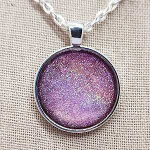 May include: A silver-toned necklace with a round pendant. The pendant features a shimmering, glittery purple design. The chain is a simple link style, and the pendant is set in a silver bezel.