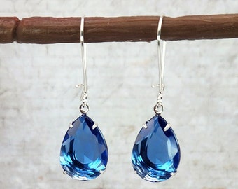 May include: A pair of elegant drop earrings featuring large, faceted, teardrop-shaped blue crystals. The crystals are set in silver-colored metal and hang from long, slender ear wires. The earrings are displayed against a neutral background.