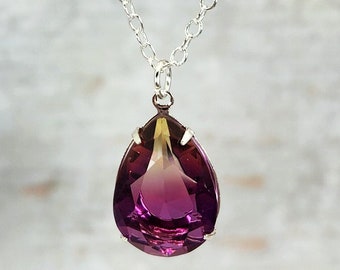 May include: A silver chain necklace with a large, teardrop-shaped pendant. The pendant features a faceted, gradient gemstone, transitioning from a deep purple to a lighter, golden hue. The gemstone is set in a silver bezel.