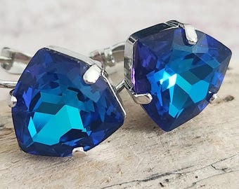 May include: A pair of blue crystal earrings. Each earring features a square-cut, faceted blue crystal set in a silver-tone metal frame. The crystals have a gradient effect, transitioning from a darker blue to a lighter, teal hue. The earrings are displayed on a piece of driftwood.