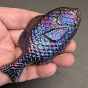 May include: A fish-shaped object with a textured, iridescent surface that shifts between deep shades of blue, purple, and dark metallic tones. The surface features a detailed scale pattern across the body and defined fins, creating a reflective appearance.