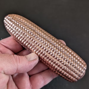 May include: A metallic, copper-colored decorative object shaped like an ear of corn. The surface features a repeating, textured pattern of small, raised kernels covering the entire cylindrical form, reflecting light with a warm, polished finish.